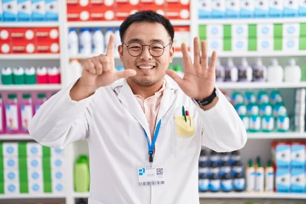 Chinese young man working at pharmacy drugstore showing and pointing up with fingers number seven while smiling confident and happy.