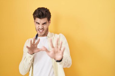 Young hispanic man standing over yellow background disgusted expression, displeased and fearful doing disgust face because aversion reaction. 