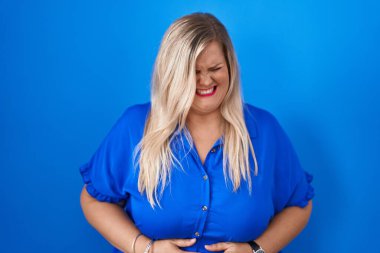 Caucasian plus size woman standing over blue background with hand on stomach because indigestion, painful illness feeling unwell. ache concept. 