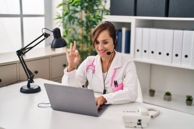 Middle age doctor woman working wearing headset doing ok sign with fingers, smiling friendly gesturing excellent symbol 