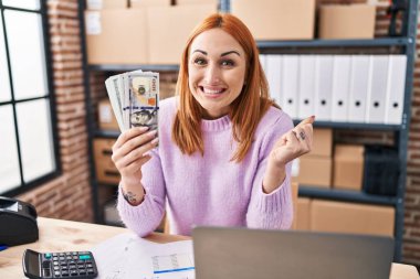 Young woman working at small business ecommerce holding money screaming proud, celebrating victory and success very excited with raised arm 