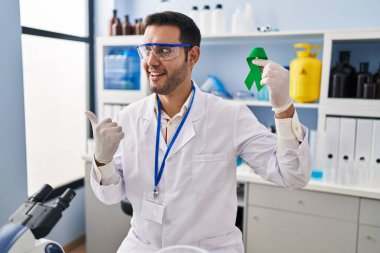 Young hispanic man with beard working at scientist laboratory holding green ribbon pointing thumb up to the side smiling happy with open mouth 