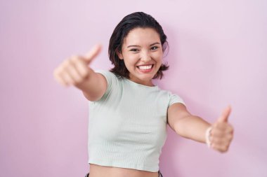 Hispanic young woman standing over pink background approving doing positive gesture with hand, thumbs up smiling and happy for success. winner gesture. 