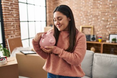 Young hispanic woman smiling confident holding piggy bank at new home