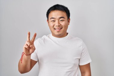 Young chinese man standing over white background showing and pointing up with fingers number two while smiling confident and happy. 