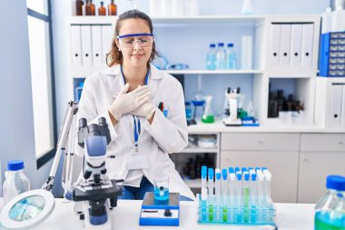 Young hispanic woman working at scientist laboratory smiling with hands on chest with closed eyes and grateful gesture on face. health concept. 