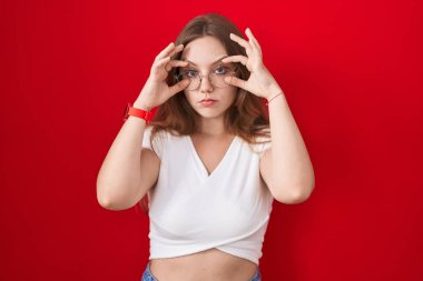 Genç beyaz kadın kırmızı arka planda duruyor parmaklarıyla gözlerini açmaya çalışıyor, uykulu ve yorgun bir şekilde sabah yorgunluğu çekiyor. 