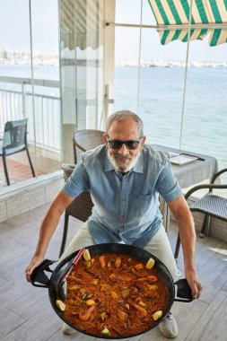 Senior grey-haired man smiling confident holding seafood paella at coffee shop terrace