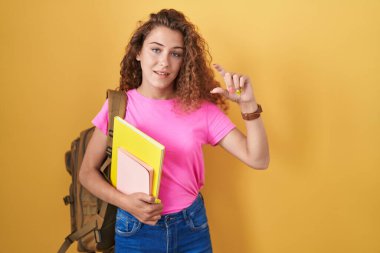 Young caucasian woman wearing student backpack and holding books smiling and confident gesturing with hand doing small size sign with fingers looking and the camera. measure concept. 