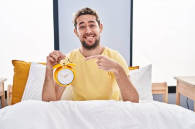 Hispanic man with beard holding alarm clock in the bed smiling happy pointing with hand and finger 