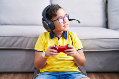 Young hispanic kid playing video game holding controller wearing headphones looking away to side with smile on face, natural expression. laughing confident. 