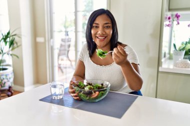 Hispanic brunette woman eating green salad at the kitchen