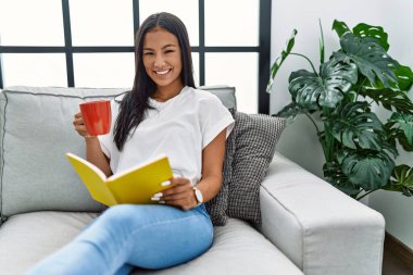 Genç Latin kadın evde kahve içip kitap okuyor.