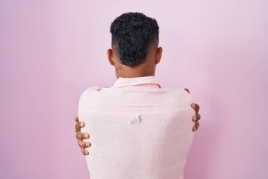 Young hispanic man standing over pink background hugging oneself happy and positive from backwards. self love and self care 