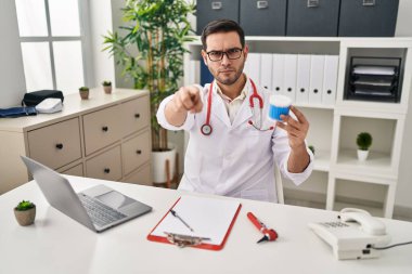 Young hispanic doctor man with beard holding ear cotton buds pointing with finger to the camera and to you, confident gesture looking serious 