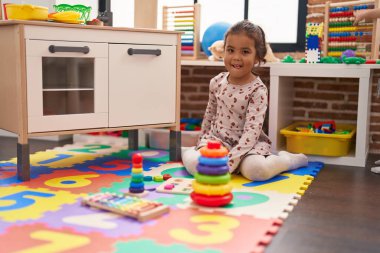 Adorable hispanic girl playing with maths puzzle game sitting on floor at kindergarten