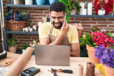 Young arab man florist talking on smartphone using laptop at florist