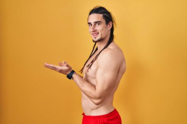Hispanic man with long hair standing shirtless over yellow background pointing aside with hands open palms showing copy space, presenting advertisement smiling excited happy 
