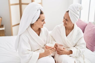 Two women mother and daughter wearing bathrobe drinking coffee at bedroom