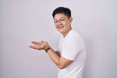 Young asian man standing over white background pointing aside with hands open palms showing copy space, presenting advertisement smiling excited happy 