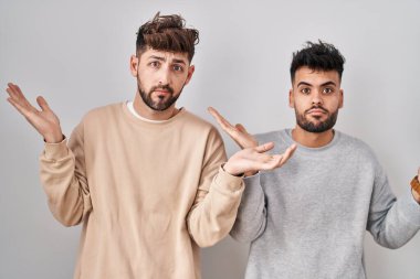Young homosexual couple standing over white background clueless and confused expression with arms and hands raised. doubt concept. 
