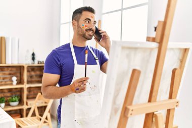 Young hispanic man smiling confident talking on the smartphone drawing at art studio