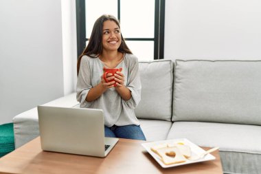 Genç Latin kadın kahvaltısını yapıyor. Evdeki kanepede oturuyor.