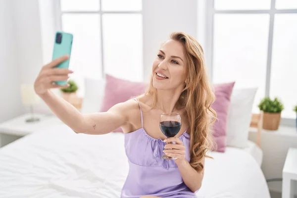 Young blonde woman making selfie by the smartphone drinking wine at bedroom