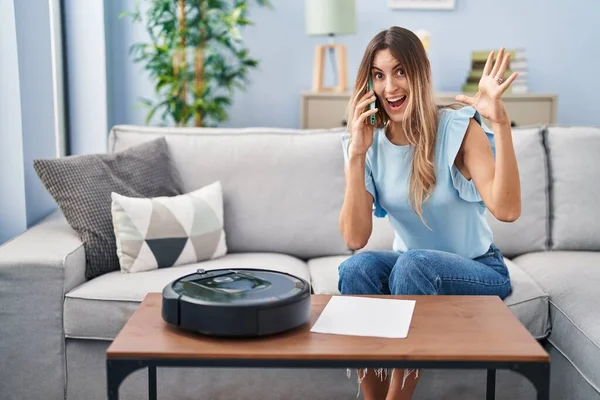 Young Hispanic Woman Sitting Home Vacuum Robot Speaking Phone Celebrating — Stockfoto
