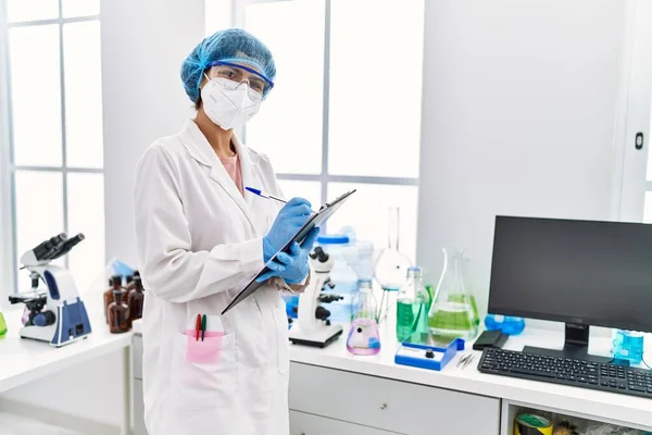实验室里穿着科学家制服在剪贴板上写字的年轻拉丁女人 — 图库照片