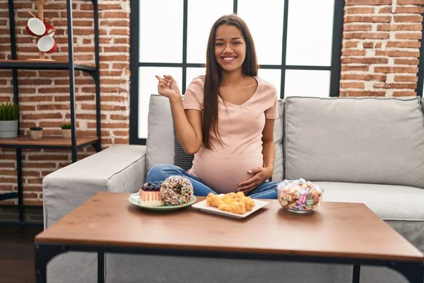 若いです妊娠中の女性食べるお菓子で自宅笑顔幸せなポインティングで手と指で側面 — ストック写真
