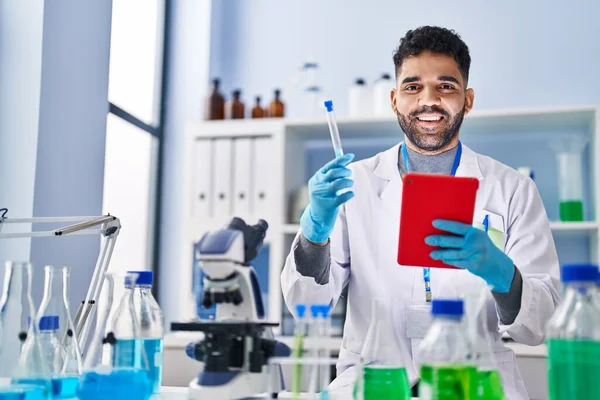 Young hispanic man wearing scientist uniform using touchpad holding test tube at laboratory