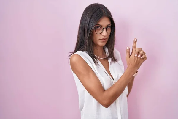 Brunette young woman standing over pink background wearing glasses holding symbolic gun with hand gesture, playing killing shooting weapons, angry face