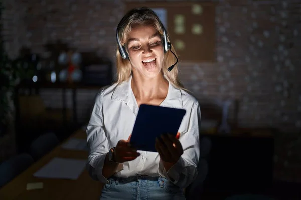 Young blonde woman working at the office at night sticking tongue out happy with funny expression. emotion concept.