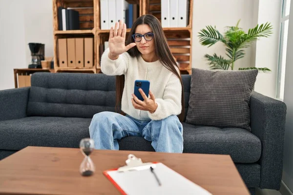 Young hispanic woman going to therapy at consultation office with open hand doing stop sign with serious and confident expression, defense gesture