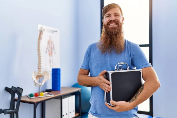 Young redhead man wearing physiotherapist uniform holding briefcase at physiotherapy clinic