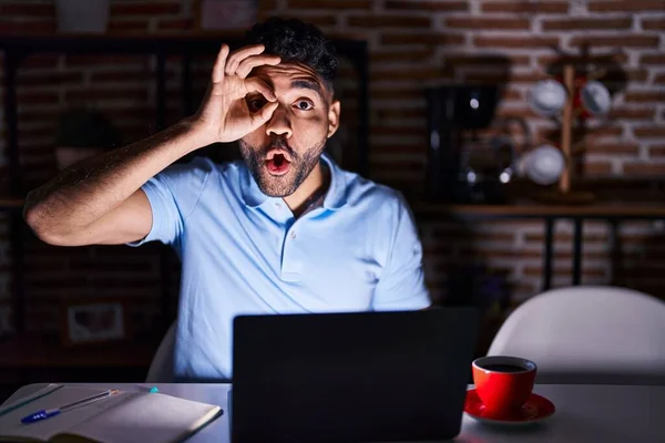 Hispanic man with beard using laptop at night doing ok gesture shocked with surprised face, eye looking through fingers. unbelieving expression.