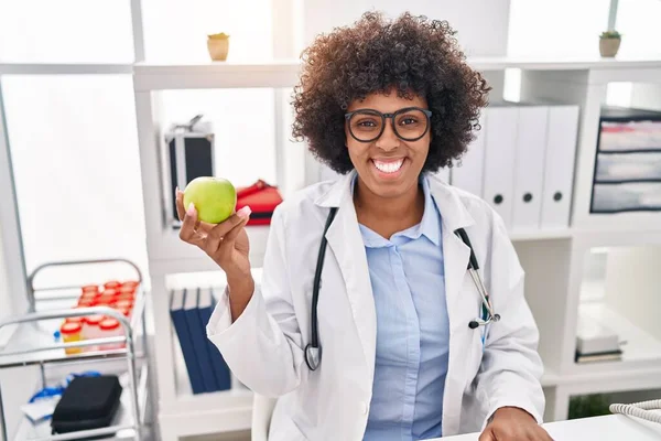 Black doctor woman with curly hair holding green apple looking positive and happy standing and smiling with a confident smile showing teeth