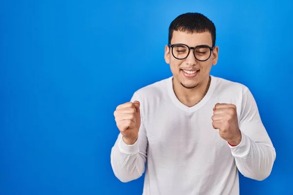 年轻的阿拉伯男子身穿休闲装 戴着眼镜 非常高兴而兴奋地举起双臂做着获胜的手势 微笑着 尖叫着要成功 庆祝概念 — 图库照片