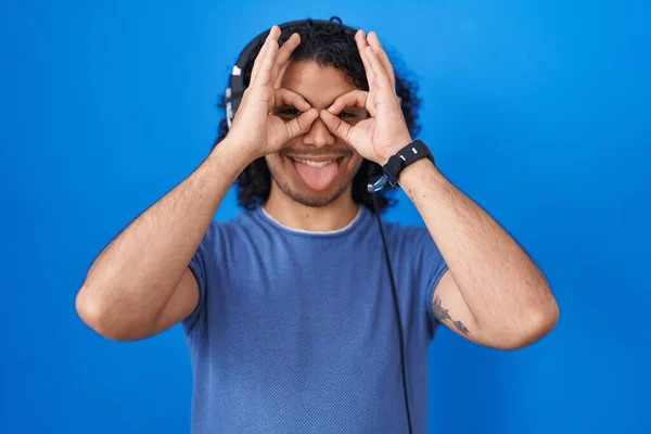 西班牙裔男人 用耳机听音乐 像双筒望远镜一样做手势 伸出舌头 眼睛透过手指看东西 疯狂的表达 — 图库照片