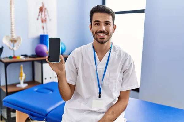 Young Hispanic Physiotherapist Man Holding Smartphone Clinic Looking Positive Happy — Stockfoto