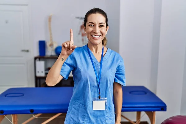 身穿理疗制服的年轻惊慌失措的女人站在诊所里 用第一指尖指尖 自信而快乐地微笑着 — 图库照片