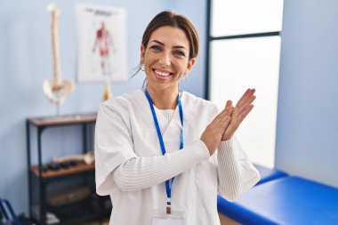 Young brunette woman working at pain recovery clinic clapping and applauding happy and joyful, smiling proud hands together 
