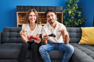 Evde video oyunu oynayan genç bir çift ellerinde kumandayla mutlu bir şekilde el ve parmak işareti yapıyor. 