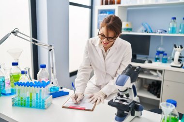 Genç beyaz kadın bilim adamı üniforması giyiyor. Laboratuvarda panoya yazı yazıyor.