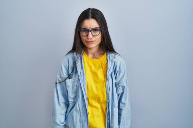 Mavi arka planda duran Latin kökenli genç kadın şüpheci ve gergin, sorun yüzünden surat asıyor. negatif kişi. 