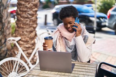 Afro-Amerikalı kadın bilgisayarını kullanıp kahve dükkanının terasında oturmuş akıllı telefondan konuşuyor.