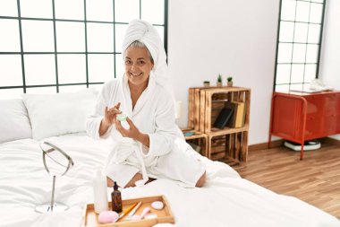 Yatak odasındaki yatakta oturan, yüz kremi kullanan orta yaşlı, kır saçlı bir kadın..