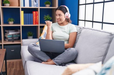 Hispanic young woman using laptop at home wearing headphones smiling with happy face looking and pointing to the side with thumb up. 