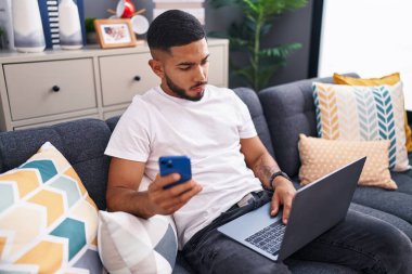Young latin man using smartphone and laptop sitting on sofa at home